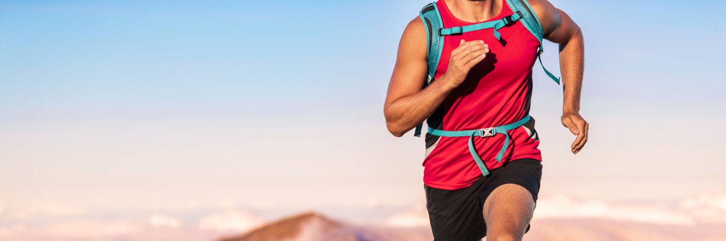 Athlete runner man running fast with backpack outdoors on blue sunset sky background with copy space. Banner panorama. Crop of midsection body, torso and arms.